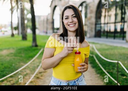 Lächelnde junge Frau mit Orangensaftflasche, während sie im Park steht Stockfoto