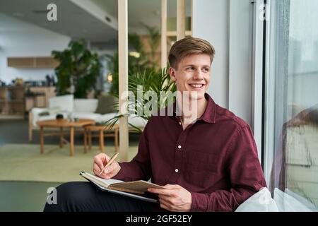 Glücklicher junger Geschäftsmann, der mit einem Tagebuch im Büro dasitzt und wegschaut Stockfoto