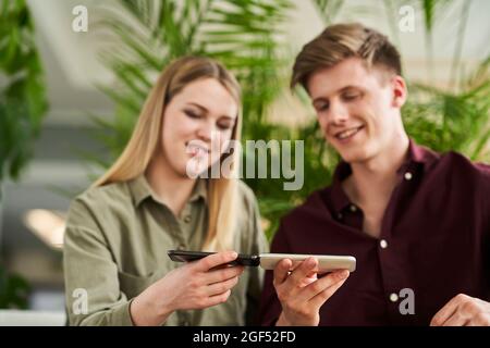 Weibliche Auszubildende im Teenageralter mit einem professionellen Mann, der im Büro die Ladeteilungstechnologie über Mobiltelefone nutzt Stockfoto