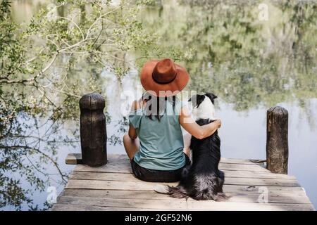 Frau mit Hut sitzend mit Arm um Haustier Hund am Pier am See Stockfoto