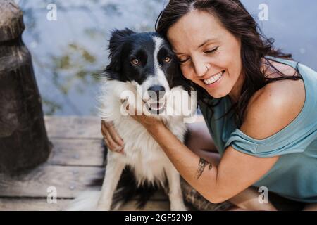 Lächelnde Frau mit geschlossenen Augen umarmt den Hund, während sie auf dem Pier sitzt Stockfoto