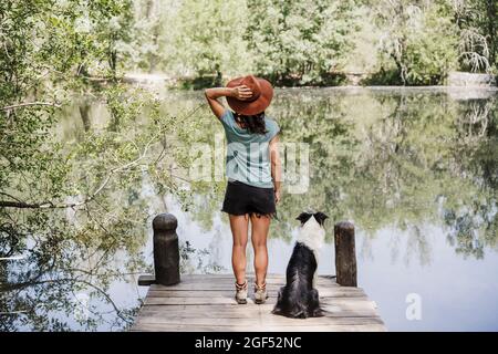 Frau mit mittlerem Erwachsenen im Hut, die am Pier am See im Wald neben einem Hund steht Stockfoto