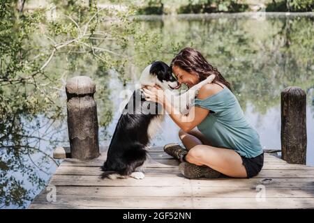 Lächelnde Frau, die Hund umarmt, während sie am Pier am See im Wald sitzt Stockfoto