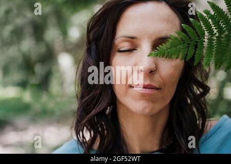 Frau mit geschlossenen Augen hält Farnblatt im Wald Stockfoto