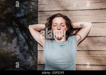 Entspannte Frau mit Händen hinter dem Kopf, die auf dem Pier am See im Wald liegt Stockfoto