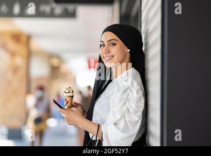 Lächelnde Frau im Hijab, die ihr Mobiltelefon hielt, während sie Eis aß Stockfoto