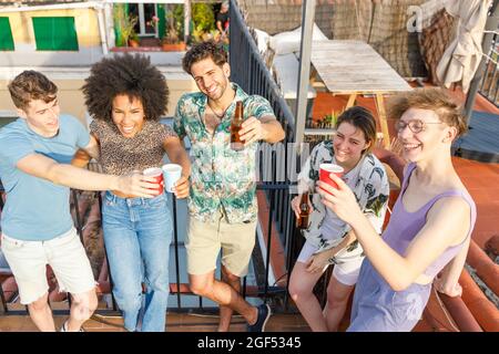 Fröhliche, multiethnische Freunde, die während der Party auf dem Dach Getränke genießen Stockfoto
