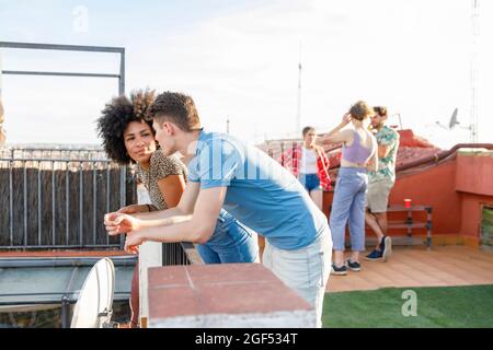 Junges Paar, das während einer Terrassenparty am Geländer angelehnt redet Stockfoto