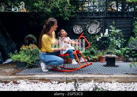 Mutter schaut auf Tochter, die auf einem Schaukelpferd im Hinterhof spielt Stockfoto