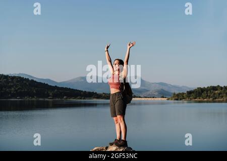 Lächelnde Wandererin mit erhobenen Armen, die am See auf einem Felsen stehen Stockfoto