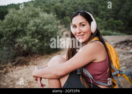 Lächelnde Frau, die über kabellose Kopfhörer Musik hört, während sie im Wald unterwegs ist Stockfoto