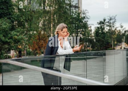 Reife Geschäftsfrau mit Smartphone, während sie sich auf Glasgeländer stützt Stockfoto