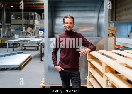 Männlicher Profi mit Hand in der Tasche, der an einem Stapel von Holzpaletten steht Stockfoto