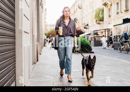 Frau mit Getränk, während sie mit einem deutschen Schäferhund auf einem Fußweg in der Stadt spazieren geht Stockfoto