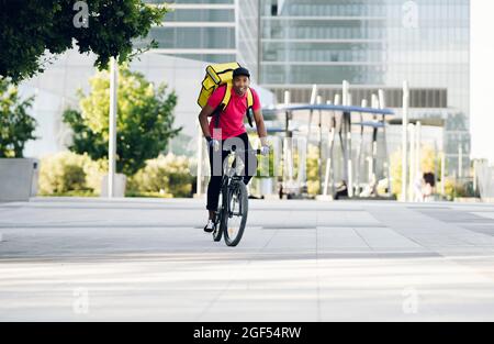 Lächelnder Lieferer mit gelbem Rucksack, der in der Stadt Fahrrad fährt Stockfoto