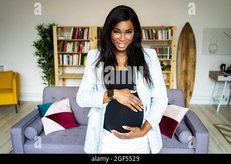 Lächelnde junge, schwanger Geschäftsfrau berührt den Bauch im Wohnzimmer zu Hause Stockfoto