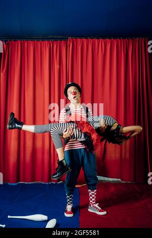 Männlicher Clown mit offenem Mund, der weibliche Darstellerin auf der Bühne im Zirkus trägt Stockfoto