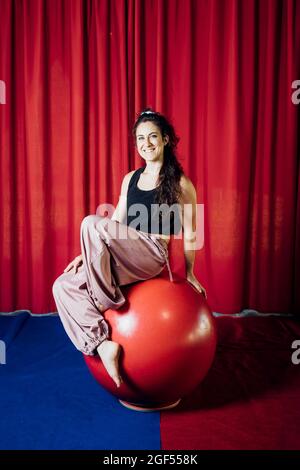 Lächelnde akrobatin, die im Zirkus auf einer roten Kugel sitzt Stockfoto