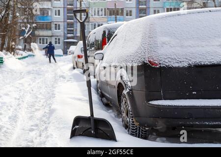 Schneeschaufel aus Kunststoff vor dem schneebedeckten Auto am sonnigen Wintermorgen mit einem Mann, der vor verschwommenem Hintergrund schaufelt Stockfoto