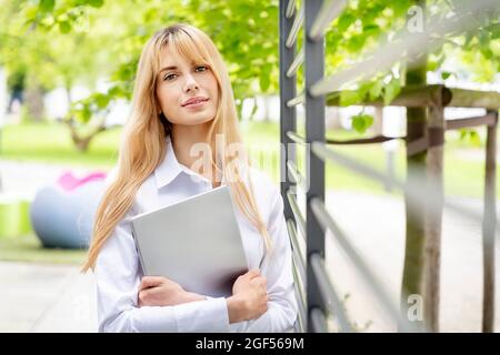 Junge blonde Geschäftsfrau hält Laptop an Metall im Büropark Stockfoto