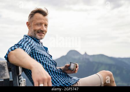 Männlicher Wanderer, der die Tasse hält, während er auf der Bank sitzt Stockfoto