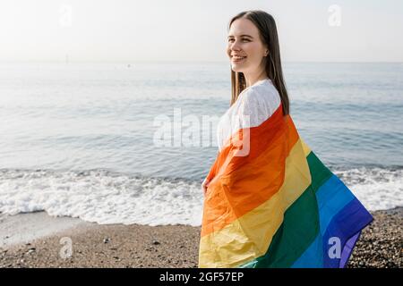 Lächelnde Frau eingewickelt in Regenbogenfahne stehend Strand Stockfoto