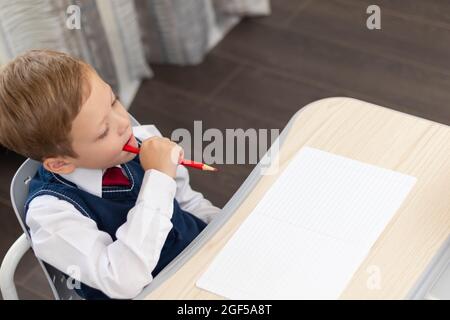 Ein süßer Erstklässler in einer Schuluniform macht Hausaufgaben, während er zu Hause mit einem Bleistift an einem Schreibtisch sitzt. Fernschulung. Selektiver Fokus Stockfoto