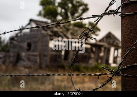 Horizontale Landschaft eines alten, verlassenen, verfaulenden und herunterfallenden Hauses im Hintergrund unscharf mit einem Stacheldrahtzaun vor der Tür Stockfoto