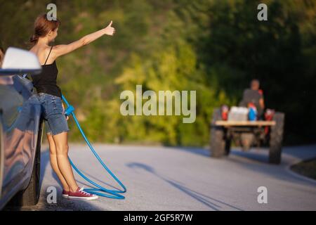 Mädchen, das neben einem Elektroauto steht und ein Ladekabel hält. Anhalter am Traktor vorbei. Stockfoto