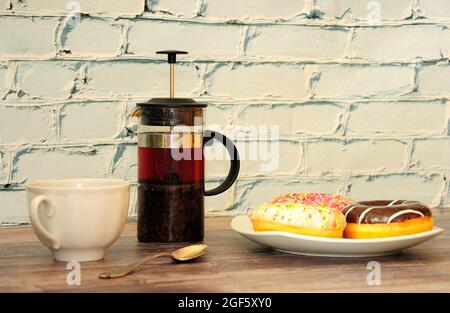 Eine Teekannen, eine Tasse schwarzen Tee, ein Löffel und ein Teller mit drei Donuts in farbenfroher Glasur stehen auf einem Holztisch an einer Ziegelwand. Nahaufnahme. Stockfoto