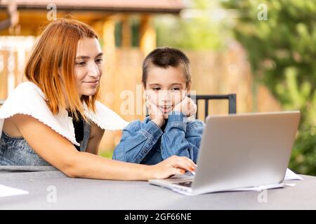 Mutter lehrt das Kind zu Hause im Garten, zu Hause Schulbildung. Mama hilft ihrem Sohn, seine Hausaufgaben zu machen, Stress. Online-Schulunterricht über Laptop Stockfoto