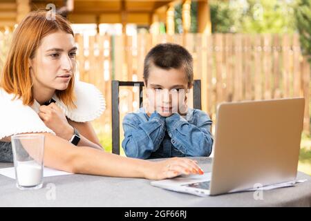 Mutter lehrt das Kind zu Hause im Garten, zu Hause Schulbildung. Mama hilft ihrem Sohn, seine Hausaufgaben zu machen, Stress. Online-Schulunterricht über Laptop Stockfoto