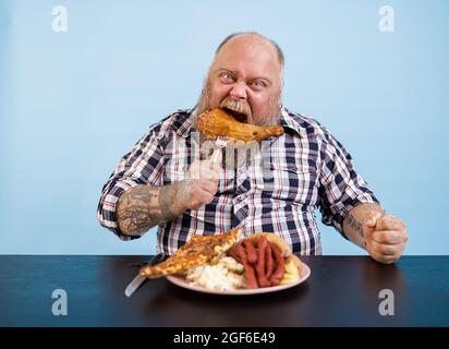 Lustiger Mann mit Übergewicht isst geräuchertes Hühnerbein am Tisch auf hellblauem Hintergrund Stockfoto