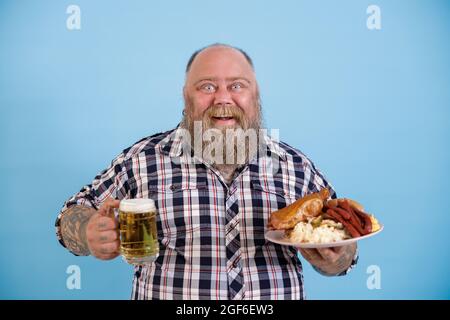 Bärtiger Mann mit Übergewicht hält fettiges Essen und Bier auf hellblauem Hintergrund Stockfoto
