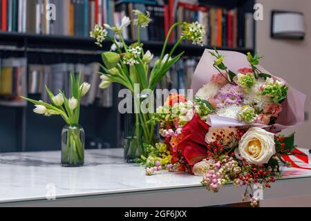 Drei wunderschöne Frühlingssträuße. Blumenarrangements mit verschiedenen Farben in Glasvase auf weißem Tisch. Unscharfer Hintergrund mit Bücherregalen Stockfoto