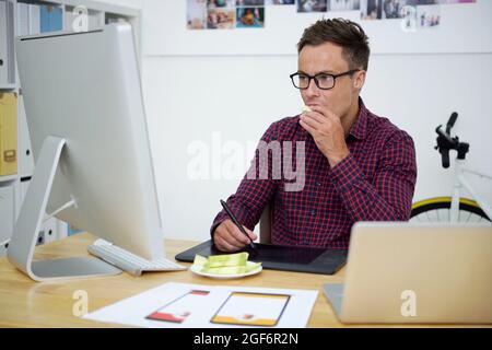 Konzentrierter junger Grafikdesigner, der am Computer arbeitet und am Schreibtisch geschnittene Früchte isst Stockfoto