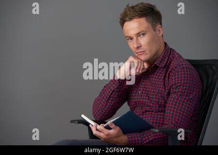 Gut aussehend für junge Mann in karierten Hemd sitzen in Stuhl und lesen Interessantes Buch Stockfoto