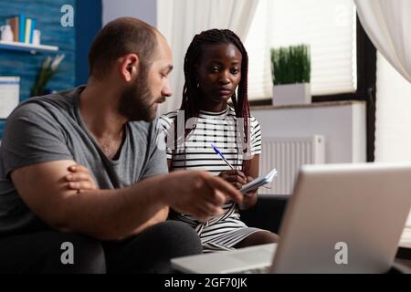 Junges interrassisches Paar, das zuhause Steuern für die Finanzwirtschaft zahlt. Multiethnische Menschen, die mit einem Laptop Steuerzahlungen überprüfen und mit Technologie Budgetgelder berechnen. Steuerabgaben von Partnern Stockfoto