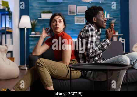 Interrassisches Paar, das wütend über Beziehung kämpft, während es auf der Couch sitzt. Multiethnische Liebhaber, die wegen Problemen und Streitigkeiten argumentieren. Frustrierte Menschen, die kämpfen Stockfoto