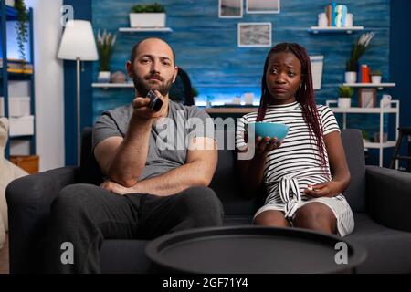 POV von interracial paar Fernsehen und entspannen, Blick auf die Kamera. Kaukasischer Mann, der die TV-Fernbedienung zum Umschalten der Kanäle verwendet, und afroamerikanische Frau, die Popcorn-Snacks hält Stockfoto