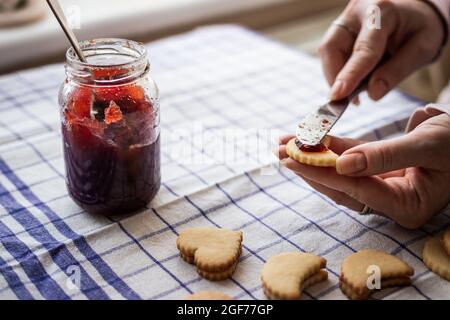 Frau, die traditionelle Weihnachtskekse von Linzer mit roter Johannisbeerkonfitüre füllt. Hausgemachter süßer Keks Stockfoto