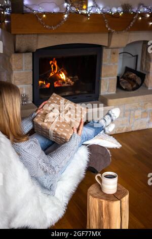 Frau mit weihnachtsgeschenk-Box und sitzt auf einem Stuhl in der Nähe des Kamins. Kaffeetasse auf Baumstumpf im gemütlichen Zuhause. Freude an den weihnachtsferien Stockfoto