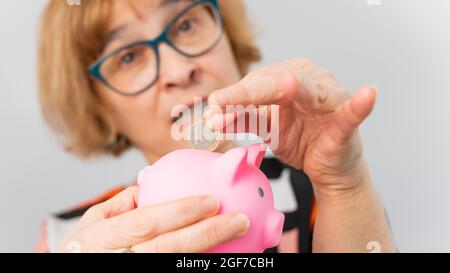 Eine ältere Frau mit Brille legt eine Münze in ein Sparschwein auf weißem Hintergrund Stockfoto