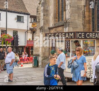 Eine Fußgängerzone an einem geschäftigen Tag. Neben einer Kirche befindet sich ein Anhänger, der Eis serviert. Zwei Männer essen Eis, während die Leute vorbei gehen. Stockfoto