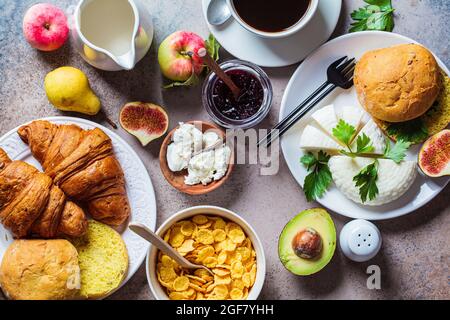 Frühstückstisch mit Cornflakes, Croissants, Obst, Käse und einer Tasse Kaffee. Dunkler Hintergrund, Draufsicht. Stockfoto