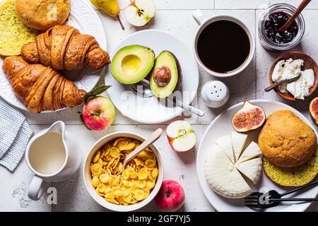 Frühstückstisch mit Cornflakes, Croissants, Obst, Käse und einer Tasse Kaffee. Grauer Kachelhintergrund, Draufsicht. Stockfoto