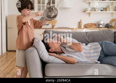 Gestresste Mutter versuchen, auf dem Sofa zu schlafen leiden unter Kopfschmerzen von frechen Sohn bang Metall Küchengeschirr Stockfoto