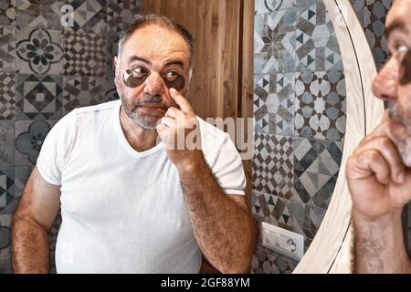Hübscher, bezaubernder, grauhaariger bärtiger Mann mit Hydrogel-Augenflecken im Gesicht. Männer Hautpflege Morgen Routine. Stockfoto