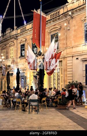 MOSTA, MALTA - 15. Aug 2021: Maltese mit Getränken, Snacks in der Open-Air-Bar. Straßendekoration von Flaggen. Covid-Vorsichtsmaßnahmen. Titularfest der Himmelfahrt Stockfoto