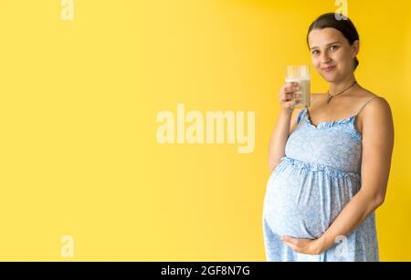 Portrait niedlich schöne schwanger junge hübsche Frau in floralen blauen Kleid hält trinken Glas Milch reibt Bauch auf gelbem Hintergrund. Mutterschaft Stockfoto